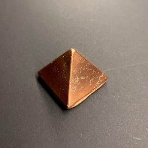 Op de afbeelding: Een kleine, koperkleurige piramide met een textuur. De vier zijden hellen naar binnen om samen te komen in een punt. Het object is decoratief, mogelijk een presse-papier of een klein beeldhouwwerk.