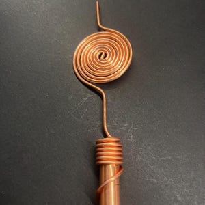 May include: A copper-colored metal object with a spiral design at the top and a cylindrical base. The spiral is a tight coil, and the base is wrapped with wire. The object is set against a dark background.