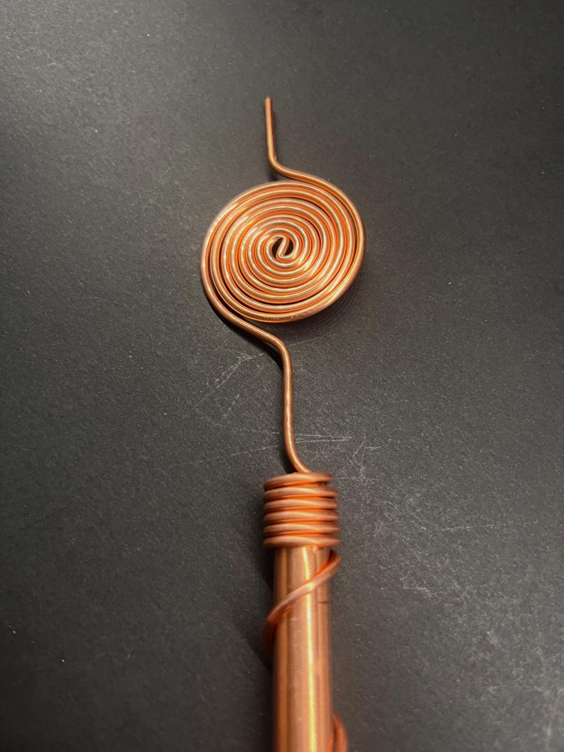 May include: A copper-colored metal object with a spiral design at the top and a cylindrical base. The spiral is a tight coil, and the base is wrapped with wire. The object is set against a dark background.