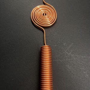 May include: A copper-colored, handmade tool with a spiral top and a coiled cylindrical handle. The top features a circular spiral design, connected to the handle by a curved wire. The handle is tightly coiled, creating a textured appearance. The tool is set against a dark background.