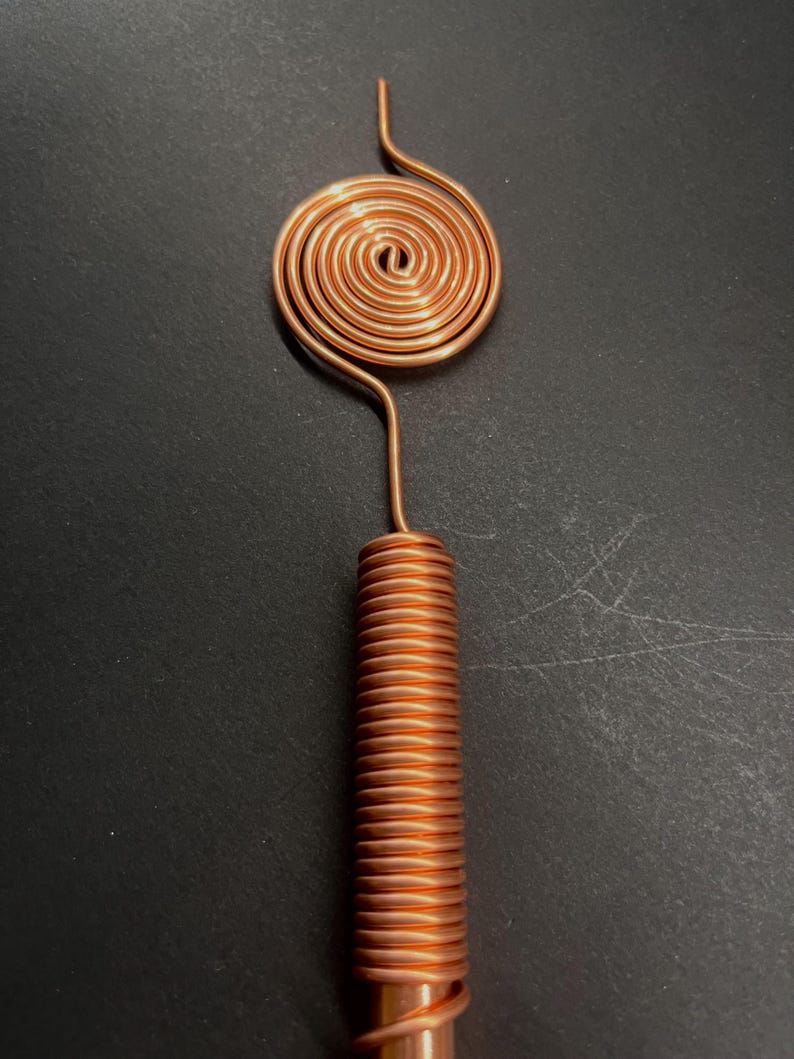 May include: A copper-colored, handmade tool with a spiral top and a coiled cylindrical handle. The top features a circular spiral design, connected to the handle by a curved wire. The handle is tightly coiled, creating a textured appearance. The tool is set against a dark background.