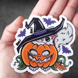 Op de afbeelding: Een witte kat met een zwarte heksenhoed met een paarse band en een witte pompoen met een jack-o'-lantern gezicht. De kat kijkt uit de bovenkant van de pompoen. Er zijn vleermuizen en het woord "Boo!" op de sticker.