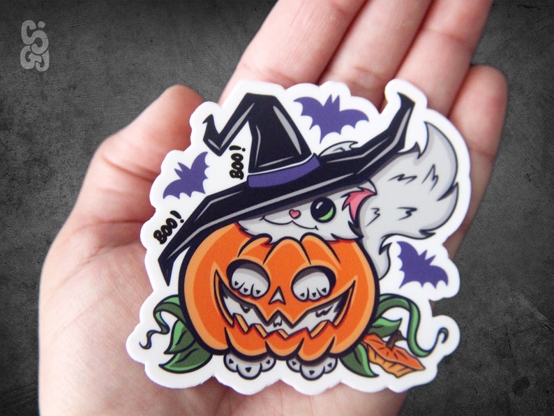 Op de afbeelding: Een witte kat met een zwarte heksenhoed met een paarse band en een witte pompoen met een jack-o'-lantern gezicht. De kat kijkt uit de bovenkant van de pompoen. Er zijn vleermuizen en het woord "Boo!" op de sticker.