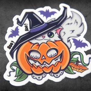 Op de afbeelding: Een witte kat met een heksenhoed zit in een uitgeholde oranje pompoen met een jack-o'-lantern gezicht. De kat heeft groene ogen en roze wangen. Er zijn vleermuizen en het woord "Boo!" op de sticker.
