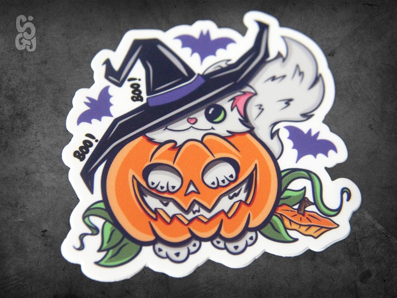 Op de afbeelding: Een witte kat met een heksenhoed zit in een uitgeholde oranje pompoen met een jack-o'-lantern gezicht. De kat heeft groene ogen en roze wangen. Er zijn vleermuizen en het woord "Boo!" op de sticker.