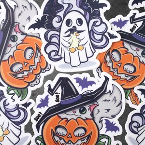 Op de afbeelding: Vier Halloween-stickers met een witte kat met een heksenhoed, een geest die een kleine gele eend vasthoudt en twee witte katten in pompoenen. De stickers hebben een witte achtergrond met zwarte vleermuizen en het woord "Boo!" op sommige van hen.