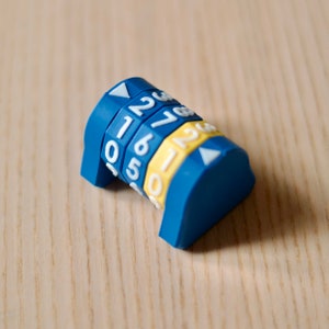 May include: A blue plastic combination lock with white numbers 1, 2, 3, 4, 5, 6, and 7. The lock has a yellow accent on each of the dials.