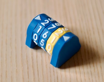 Puede incluir: Una cerradura de combinación de plástico azul con números blancos 1, 2, 3, 4, 5, 6 y 7. La cerradura tiene un acento amarillo en cada uno de los diales.