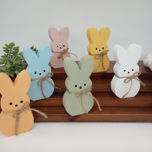 Peut inclure: Six figurines de lapin en bois dans différentes couleurs pastel, notamment bleu clair, rose, jaune, vert et blanc. Chaque lapin a un petit nœud brun noué autour de son cou. Les lapins sont disposés sur un support en bois à plusieurs niveaux.