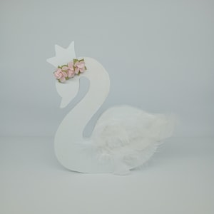 Könnte beinhalten: Weiße Holzschwanenfigur mit einer Krone und rosa Blumen auf dem Kopf. Der Schwan hat weiße Federn auf dem Rücken.