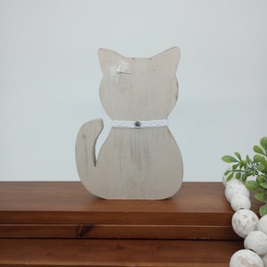 May include: A white wooden cat figurine with a white bow and a white lace collar with a blue gem in the center. The cat is sitting on a wooden shelf.