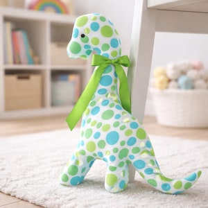 May include: A soft toy dinosaur made from white fabric patterned with green and blue polka dots. It has a green ribbon bow. The toy is on a white rug, next to a white chair.