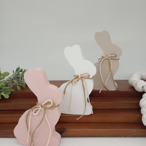Puede incluir: Tres figuritas de conejo de madera en rosa, blanco y beige, cada una con un lazo de cuerda marrón. Los conejos están sentados en una pila de estantes de madera.