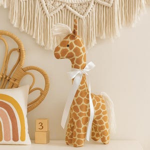 Puede incluir: Un juguete de peluche de jirafa con una cinta blanca alrededor del cuello. La jirafa tiene un patrón de manchas marrones y beige. Un tapiz de macramé blanco está en el fondo, junto con una almohada con un diseño de arcoíris y bloques de madera.