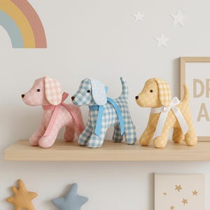 Può includere: Tre statuette di cani di peluche dai colori pastello sono su una mensola di legno. I cani sono rosa, blu e gialli, con motivi a quadretti e floreali. Ogni cane ha un nastro intorno al collo.