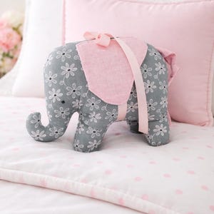 Puede incluir: Un peluche con forma de elefante gris con estampados florales blancos y una silla de montar rosa. El elefante tiene una cinta rosa y una correa. Se coloca sobre una colcha de lunares rosas y blancos. Decoración infantil.