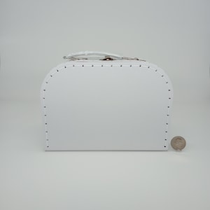 May include: A small, white, vintage-style suitcase with a rounded top and a handle. The suitcase has a metal clasp and is decorated with small, evenly spaced black stitches. A coin is next to the suitcase.