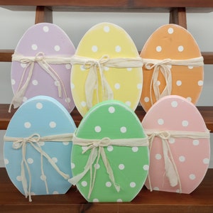 Puede incluir: Seis huevos de Pascua de madera decorados con lunares blancos y atados con una cinta blanca. Los huevos son de varios colores pastel, incluyendo azul claro, verde claro, rosa claro, amarillo claro y lavanda.