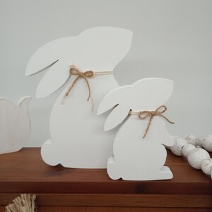 May include: Two white wooden bunny figurines, one larger than the other, sitting side-by-side on a wooden surface. Both bunnies have brown twine bows tied around their necks.