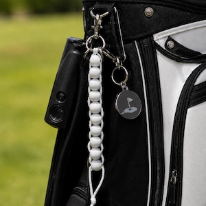 May include: A black and white golf bag with a beaded golf club counter and a golf-themed keyring. The counter is white with wooden beads. The keyring has a circular tag with a golf flag graphic.