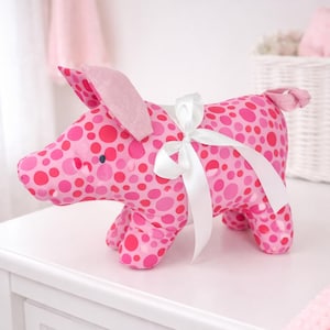 May include: A pink stuffed pig with a polka dot pattern in varying shades of pink and red. The pig has a white ribbon tied around its middle. The pig is sitting on a white surface.