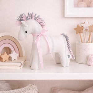 Può includere: Un morbido unicorno bianco con dettagli grigi e rosa, un nastro rosa e una coda intrecciata. L'unicorno ha una criniera grigia e rosa. Il giocattolo è su una mensola bianca con altri oggetti decorativi.