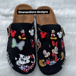 May include: A pair of black slip-on clogs with a cork platform sole. The clogs are decorated with various Mickey Mouse and Minnie Mouse patches, including a large Mickey Mouse and Minnie Mouse couple patch on each clog. The clogs also feature red and white buttons and a butterfly patch.