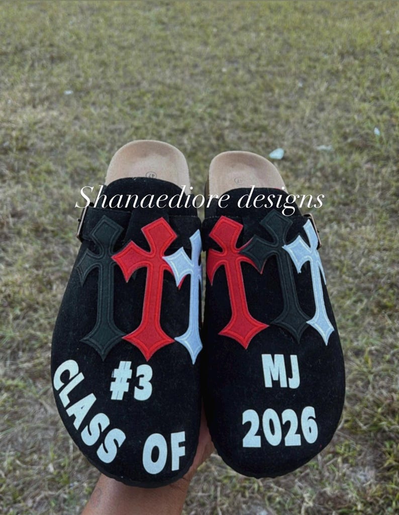May include: Black suede clogs with a white, red, and black cross design. The left clog has the text "Class of 2026 #3" and the right clog has the text "MJ 2026".