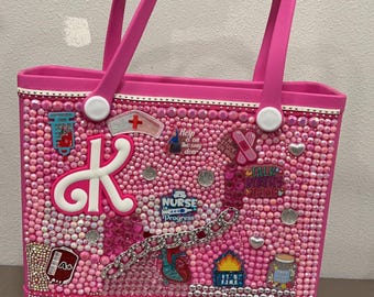 May include: A pink tote bag featuring a white letter K and a variety of colourful patches, including a nurse patch, a heart patch, and a patch that reads "Talk Pretty, Stop Being Mean". The bag is adorned with rhinestones and has a white strap. The bag is labelled "Shanaediore designs" at the bottom.