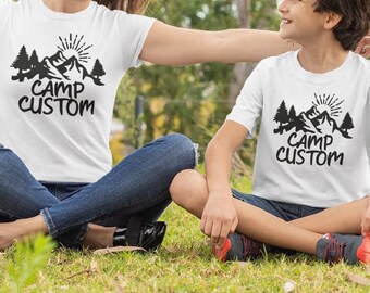 May include: A woman and a child wearing white t-shirts with a black graphic of mountains and trees and the text "Camp Custom". They are sitting on a grassy field with yellow flowers.