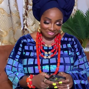 Può includere: Una donna indossa un abito blu con paillettes, una collana di perle rosse e un turbante blu. Sta tenendo uno smartphone nelle sue mani.