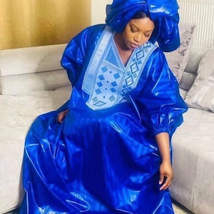 May include: A vibrant blue, flowing dress and matching head wrap. The dress features a V-neck with intricate white embroidery. The fabric has a shiny, satin-like appearance, creating a luxurious look. The sleeves are long and gathered at the wrists.