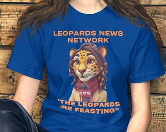 May include: A woman wearing a blue t-shirt with a cartoon leopard wearing glasses and a microphone. The shirt says "Leopards News Network" and "The Leopards Are Feasting".