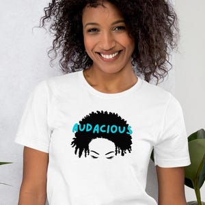 Puede incluir: Camiseta blanca con un gráfico afro negro y la palabra "AUDACIOUS" en azul. El diseño incluye una ilustración estilizada del cabello de una mujer. La camiseta está hecha de un material suave y es adecuada para ropa casual.