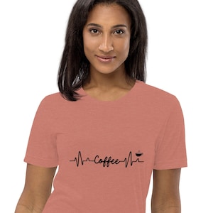 Op de afbeelding: Een heidekleurig t-shirt met een zwarte afbeelding van een hartslag met het woord "Coffee" en een kopje koffie-icoon.