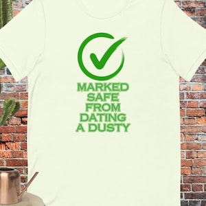 May include: Cream-colored t-shirt with a green checkmark logo and the text "MARKED SAFE FROM DATING A DUSTY" in green. The shirt is displayed on a wooden surface with a copper watering can and a cactus in the background.