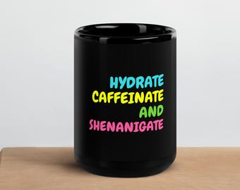 May include: A black coffee mug with the words "HYDRATE CAFFEINATE AND SHENANIGATE" in blue, yellow, and pink. The mug is on a wooden cutting board.