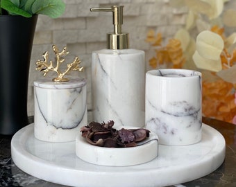 May include: A marble bathroom set with a soap dispenser, tumbler, small dish, and lidded jar, all on a round tray. The set features gold accents and a white and gray veined pattern. Decorative dried flowers and a vase of pink flowers are in the background.