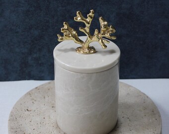 May include: A white marble container with a gold coral-shaped lid. The container is sitting on a beige stone coaster.