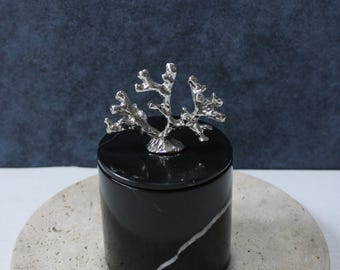 May include: Black marble jar with a silver coral design lid. The jar is sitting on a beige stone coaster.