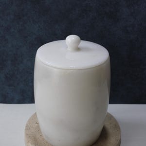 May include: White marble bathroom container with a lid. The container is sitting on a beige round coaster.