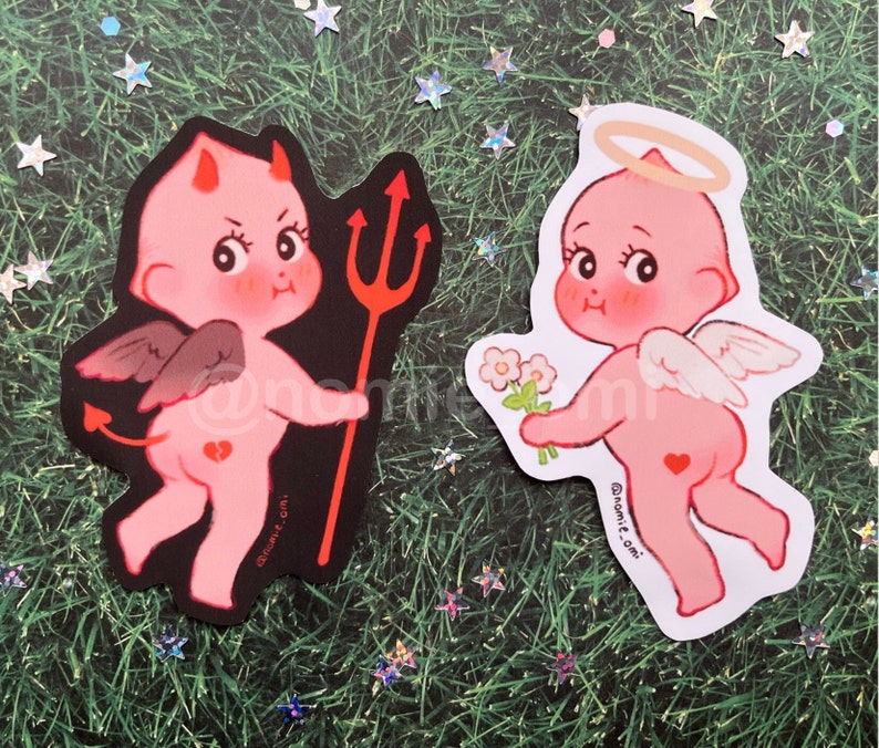 May include: Two stickers featuring cartoon cherubs. One cherub is a pink angel with a halo and wings, holding a bouquet of white flowers. The other cherub is a pink devil with horns, wings, and a tail, holding a red trident. Both stickers have a black outline and are on a green background.