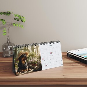 K&ouml;nnte beinhalten: Ein Tischkalender f&uuml;r den Monat Mai mit einer Spiralbindung an der Oberseite. Die linke Seite zeigt ein Foto eines schwarz-braunen Hundes mit einem Strohhut in einem Gew&auml;chshaus, w&auml;hrend die rechte Seite ein Kalenderraster f&uuml;r Mai darstellt.