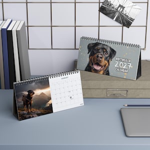 K&ouml;nnte beinhalten: Ein 2027 Rottweiler-Tischkalender mit Spiralbindung. Das Deckblatt zeigt ein Rottweiler-Portr&auml;t mit dem Text ROTTWEILER 2027 CALENDAR. Eine ge&ouml;ffnete Seite zeigt einen Rottweiler auf einem Berggipfel und ein Kalenderblatt f&uuml;r den Monat August.