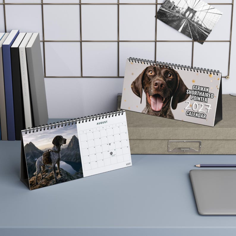 May include: A 2027 desk calendar featuring a German Shorthaired Pointer on the cover and an August page showing a dog standing on a mountain peak. The cover text reads GERMAN SHORTHAIRED POINTER 2027 CALENDAR. The calendar has a black spiral binding.