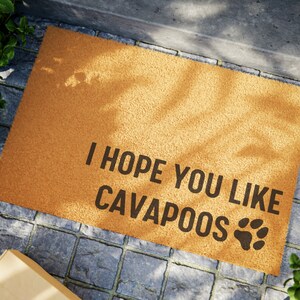 Op de afbeelding: Een rechthoekige deurmat van bruin kokosvezel. De mat heeft de zwarte tekst "I HOPE YOU LIKE CAVAPOOS" en een pootafdruk. De mat ligt op een stenen oppervlak.