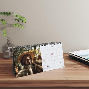 May include: A desktop spiral-bound calendar featuring a photograph of a brown dog wearing a straw hat on the left page and a white grid for the month of May on the right page. The calendar is printed on white paper with black text and wire binding.