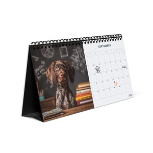 May include: A desk calendar with a black spiral binding featuring a photograph of a brown and white dog wearing glasses in front of a chalkboard with books. The right side displays the month of September with a grid and small icons on specific dates.