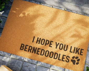 May include: A rectangular doormat in a tan color, with the text "I HOPE YOU LIKE BERNEDOODLES" in black, along with a paw print graphic. The mat is placed on a stone surface.