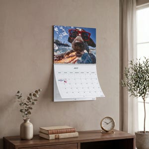 May include: A wall calendar for the month of July featuring a photograph of a brown dog wearing red sunglasses while swimming in water. The calendar grid includes a small American flag icon and the text Independence Day on the fourth of July.